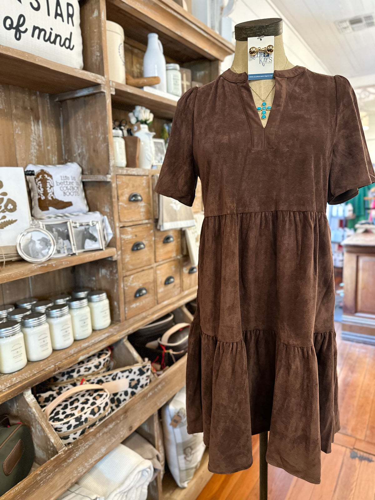 A brown short-sleeve tiered dress displayed on a mannequin in a rustic shop interior, with wooden shelves holding jars, bags, and decor in the background.