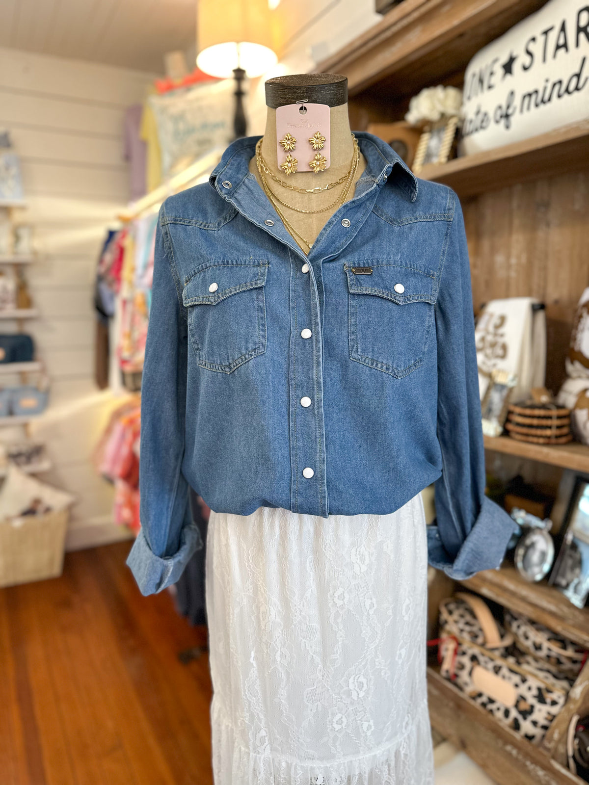 A blue denim button-up shirt with two chest pockets and white buttons, styled on a mannequin torso with a white lace skirt in a retail shop setting.