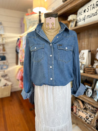 A blue denim button-up shirt with two chest pockets and white buttons, styled on a mannequin torso with a white lace skirt in a retail shop setting.