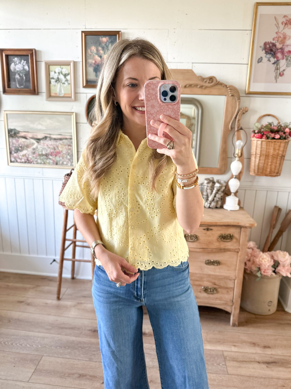 Woman taking a mirror selfie wearing a yellow lace top and blue jeans in a room with wooden furniture and decorative items.