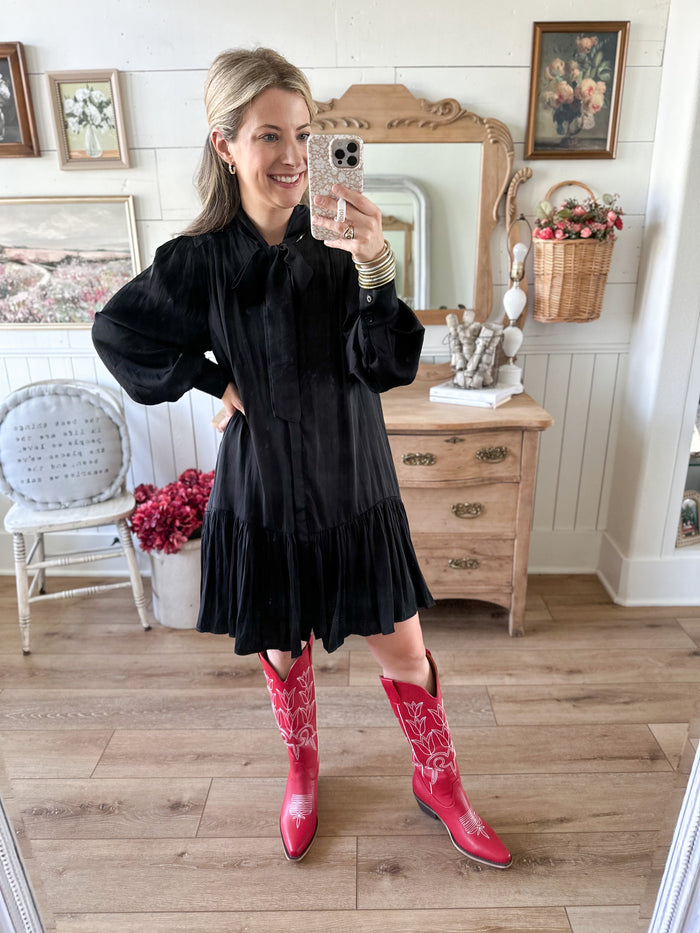 Woman taking a mirror selfie wearing a black dress and pink cowboy boots in a room with wooden floors and framed pictures on the wall.