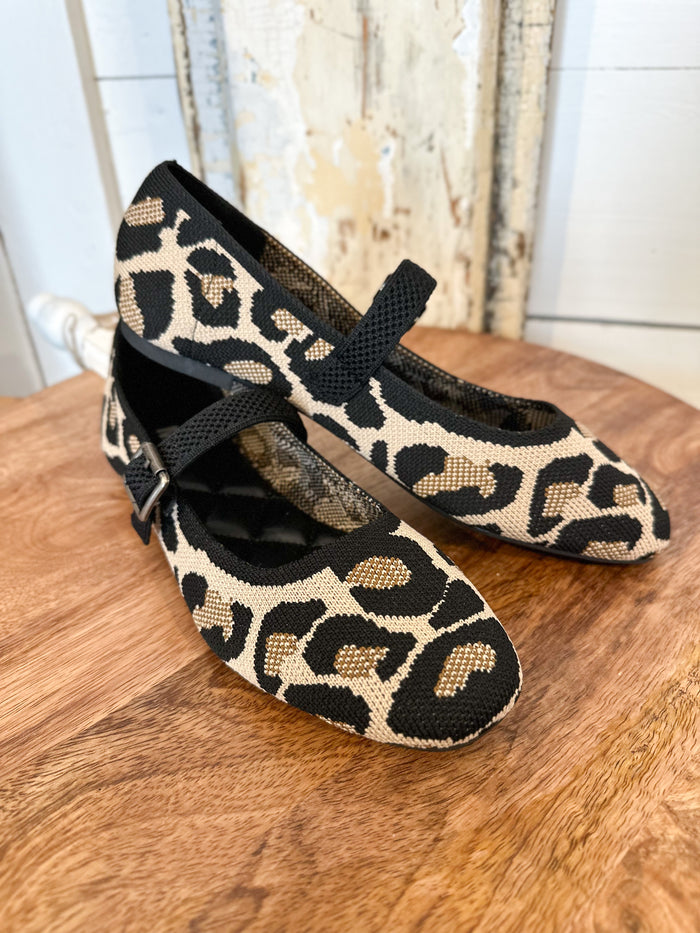 Pair of leopard print shoes with black straps on a wooden surface.