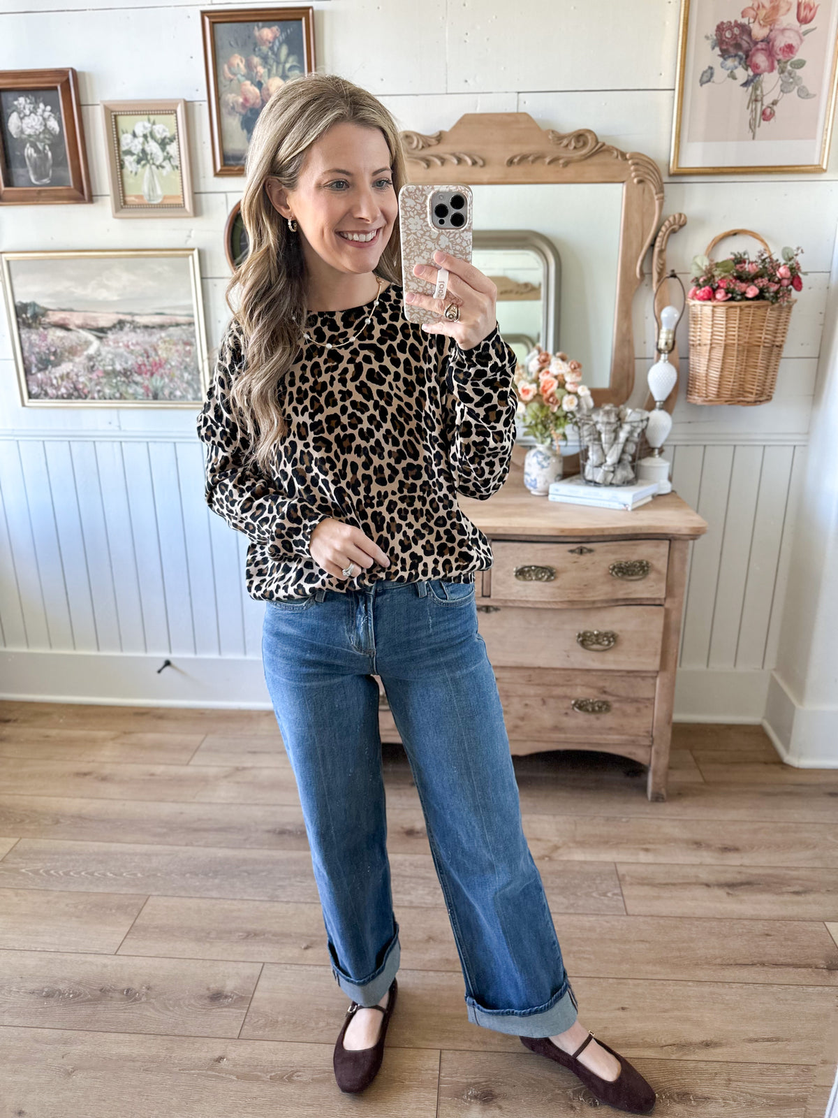 Woman taking a mirror selfie wearing a leopard print top and blue jeans in a room with wooden furniture and decorative items.