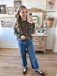 Woman taking a mirror selfie wearing a leopard print top and blue jeans in a room with wooden furniture and decorative items.