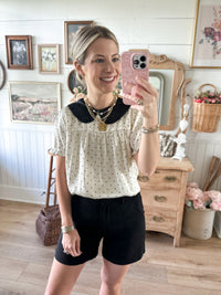 A woman takes a mirror selfie in a cozy, vintage-style room, wearing a cream short-sleeve blouse with black polka dots and a black Peter Pan collar, paired with black shorts.