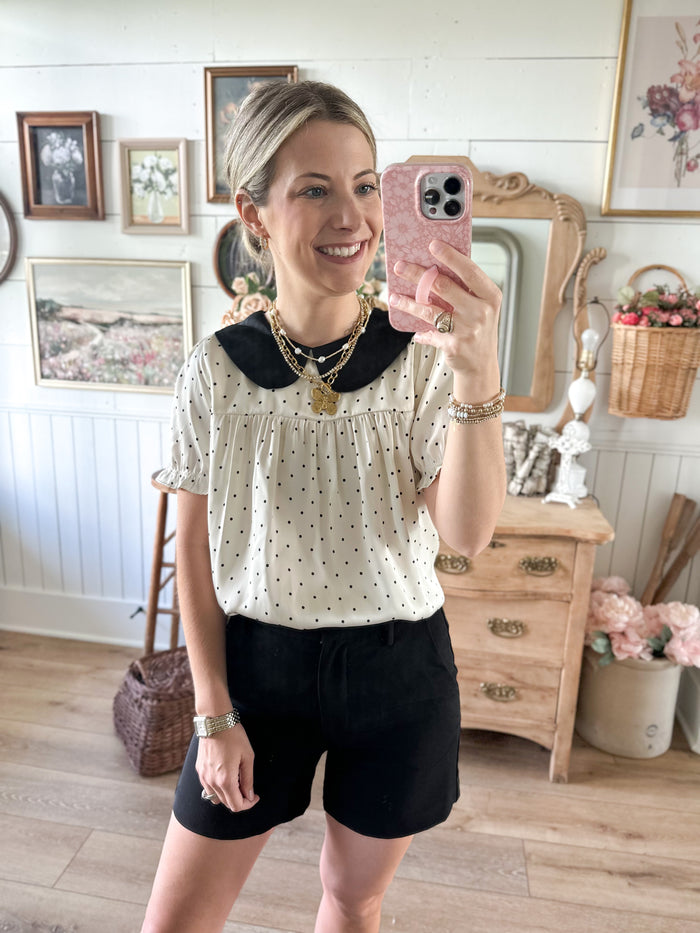 A woman takes a mirror selfie in a cozy, vintage-style room, wearing a cream short-sleeve blouse with black polka dots and a black Peter Pan collar, paired with black shorts.