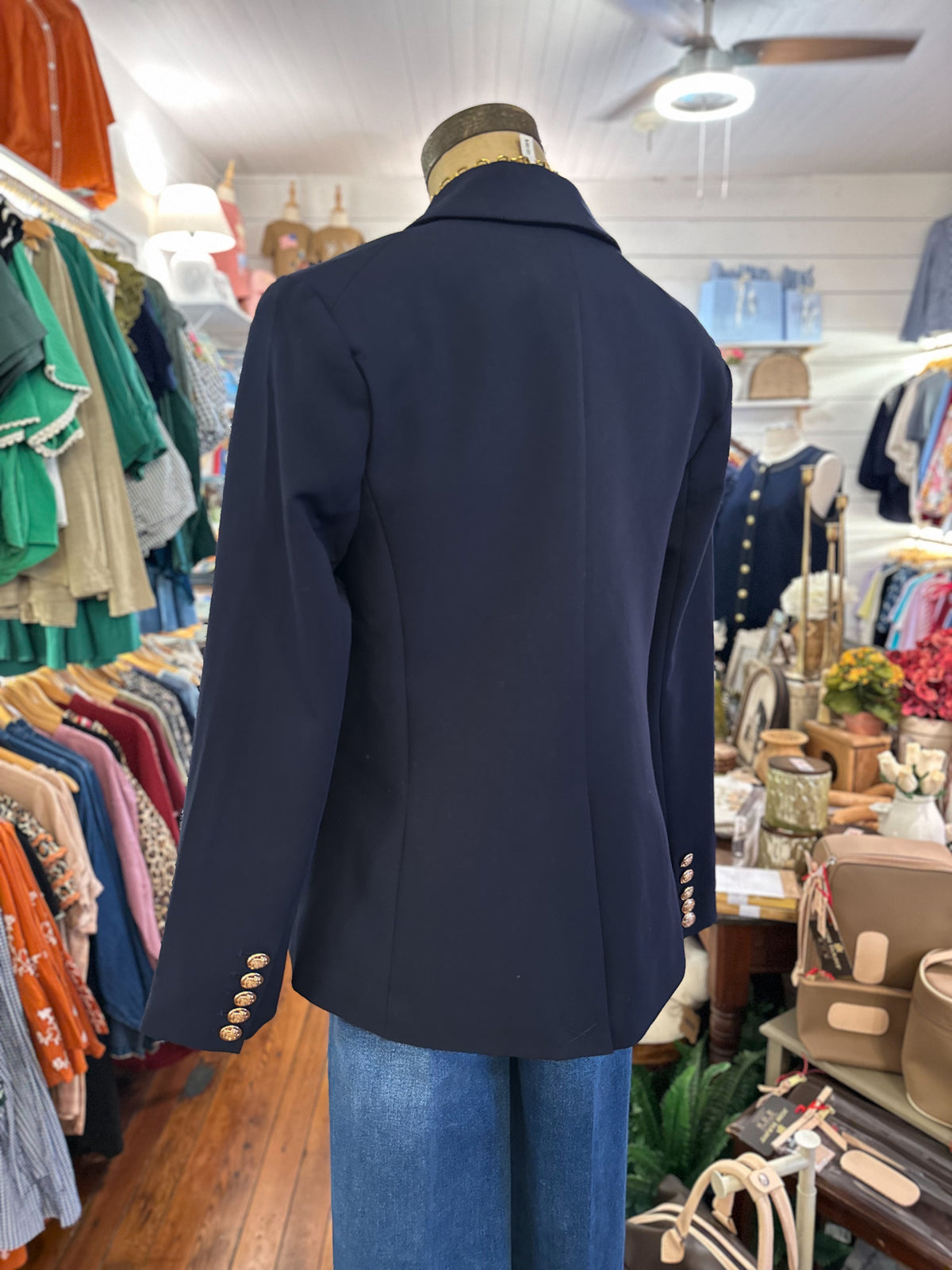 A navy blue blazer displayed on a mannequin torso in a clothing boutique, styled with blue jeans. The blazer features structured shoulders, a notched collar, and decorative gold-tone buttons on the cuffs.