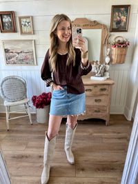 Woman taking a mirror selfie wearing a brown top, denim skirt, and white cowboy boots in a home setting.