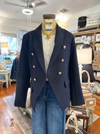 A navy blue double-breasted blazer with gold buttons displayed on a mannequin torso inside a boutique. The blazer is styled with a white top and blue jeans, with a gold necklace on the mannequin.