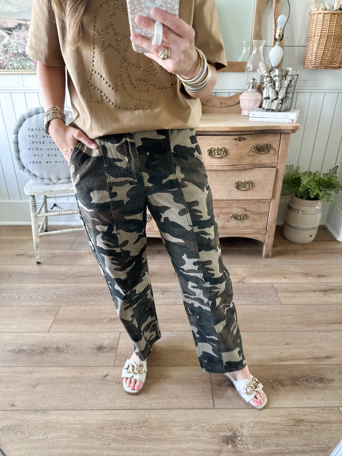 Person wearing camouflage pants and a beige top in a room with wooden flooring and furniture.