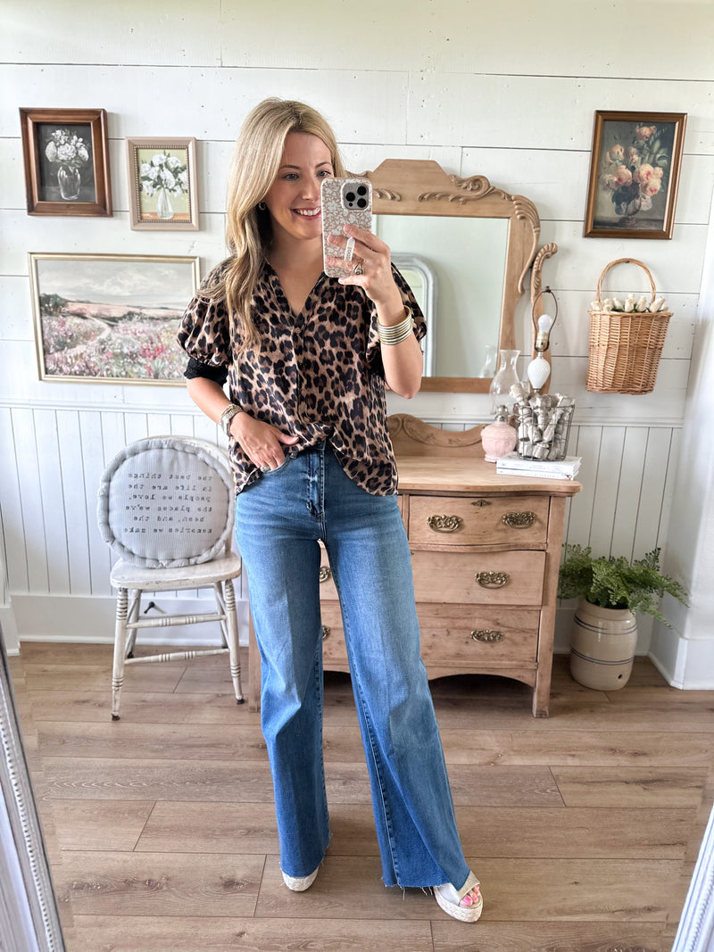 Woman taking a mirror selfie wearing a leopard print top and blue jeans in a room with wooden floors and white walls.
