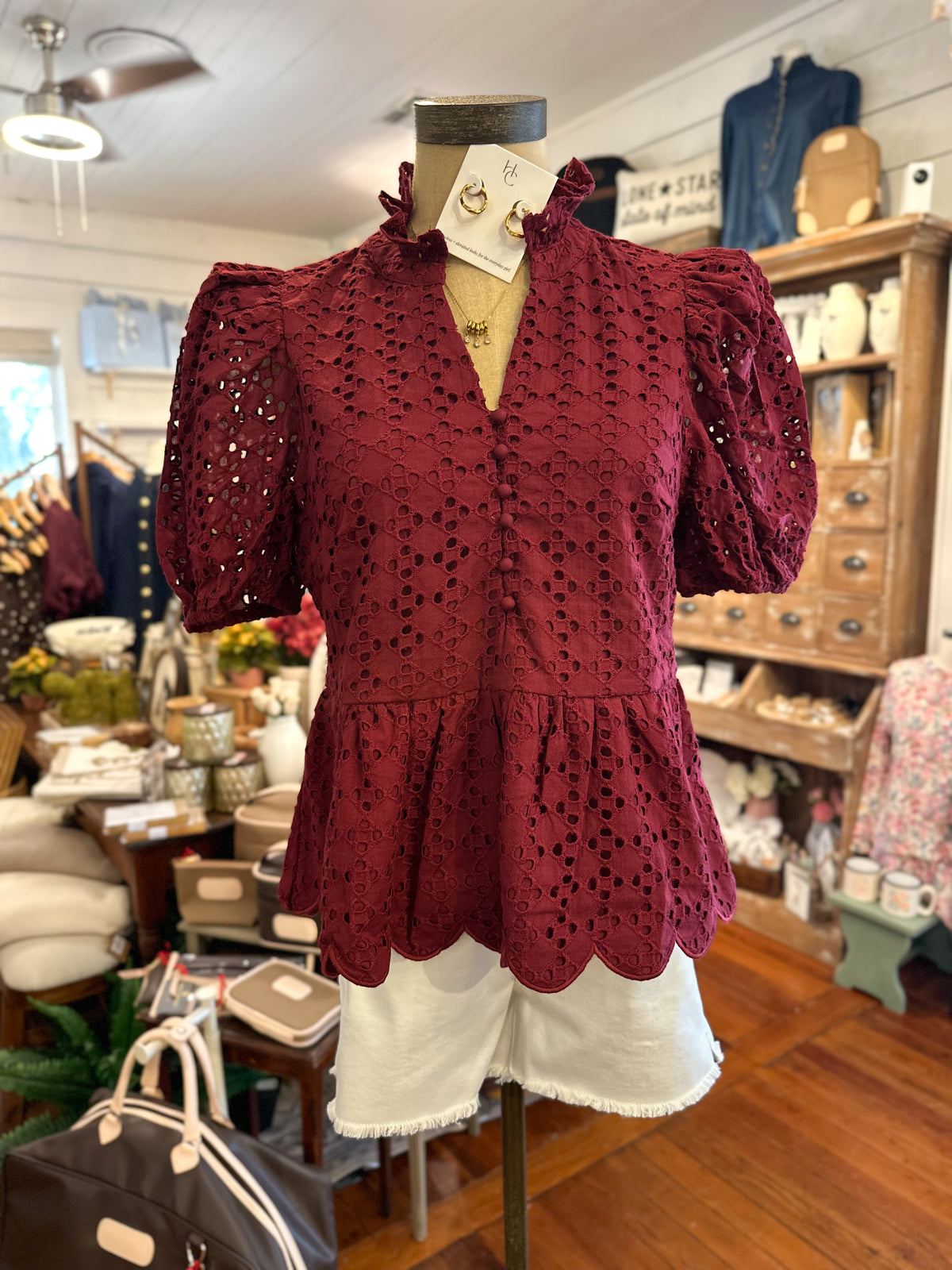 A burgundy eyelet lace blouse with puff sleeves and a ruffled mock neck, displayed on a mannequin torso with white shorts inside a boutique.