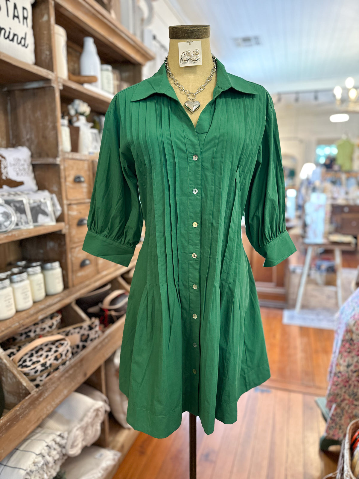A green button-front shirt dress with three-quarter sleeves and vertical pleating, displayed on a mannequin torso inside a retail store.