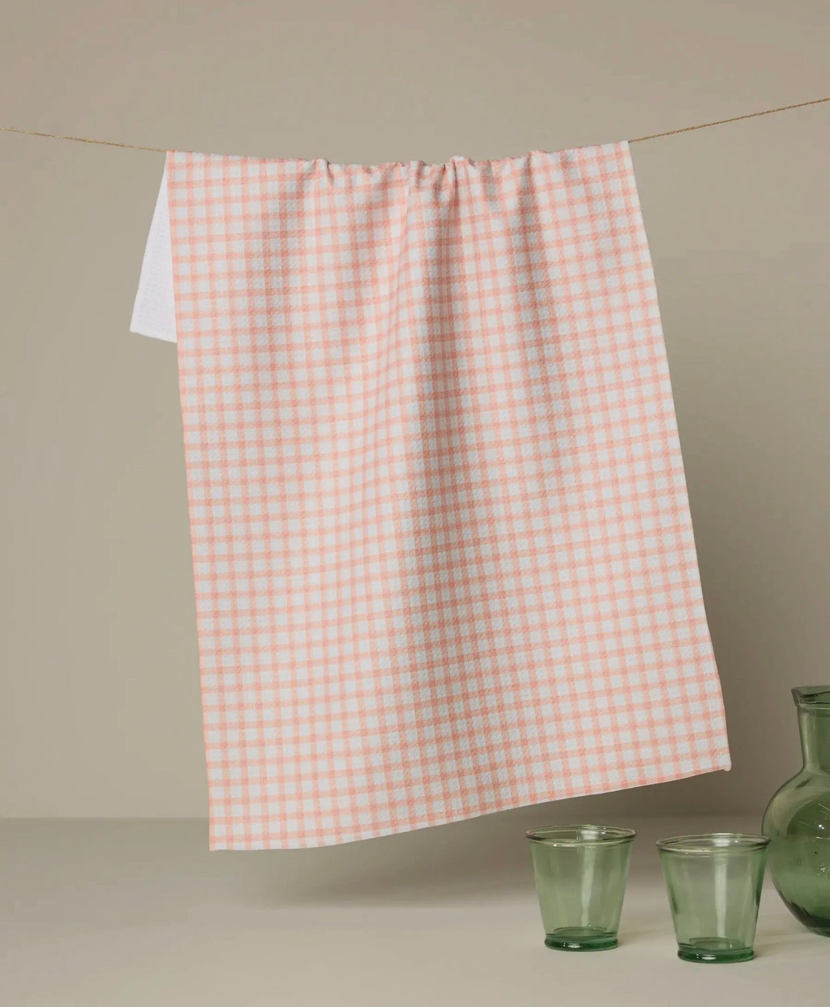 Checkered dish towel on a rod with green glasses and a vase on a neutral background peach color geometry towel 

