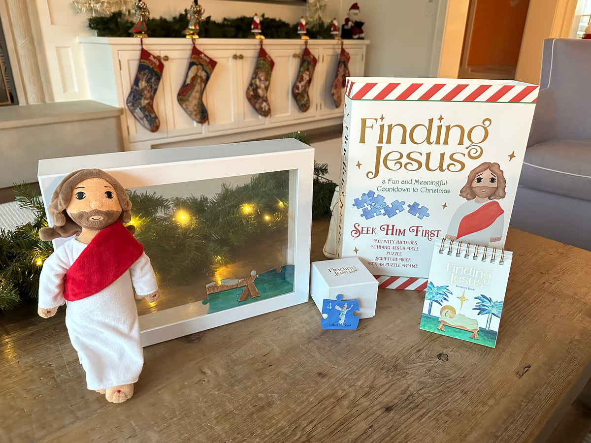 A Christmas-themed gift set on a wooden table, featuring a Jesus doll in white and red, a white shadow box with a nativity scene, a book titled 'Finding Jesus', a small white box, and a small booklet.