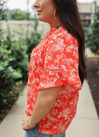 Woman wearing a red floral blouse with blurred greenery in the background
bell top layerz clothing 