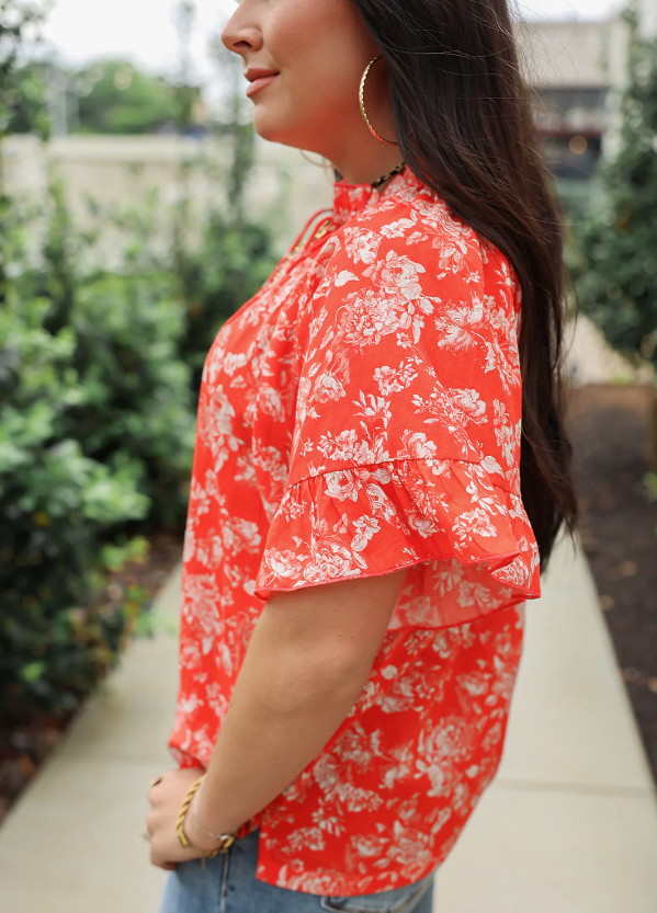 Woman wearing a red floral blouse with blurred greenery in the background
bell top layerz clothing 