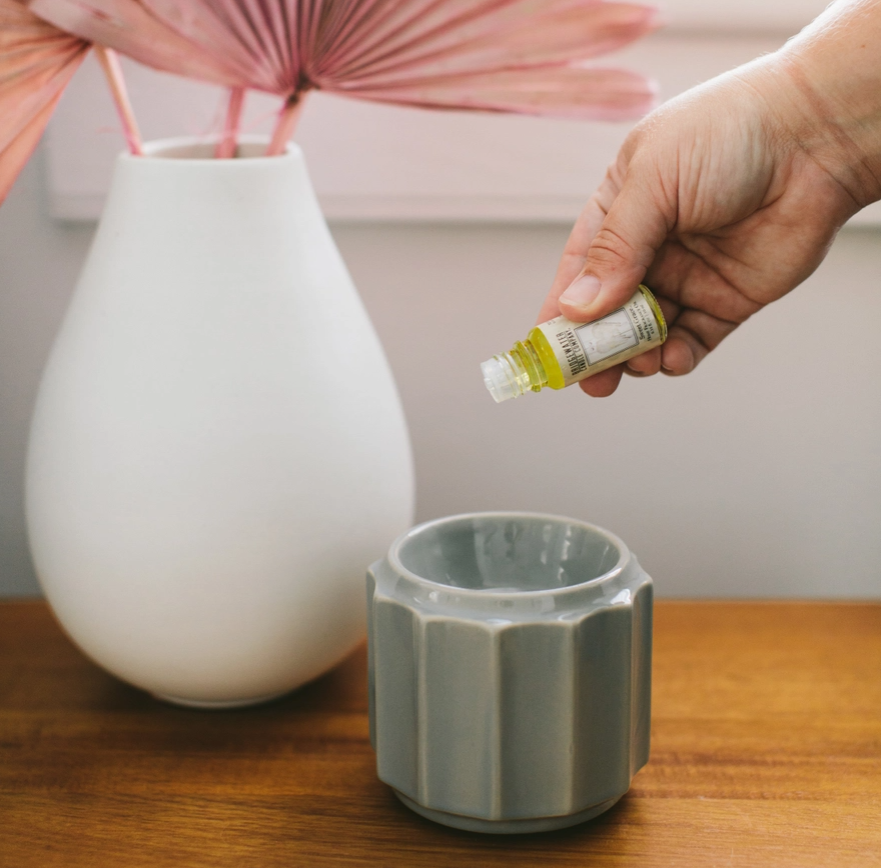 Hand holding a small bottle over a gray container on a wooden surface with a white vase and pink decorative item in the background.