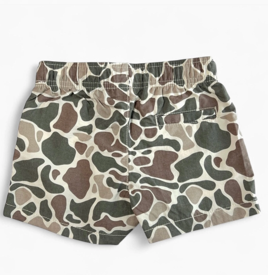 A pair of children's shorts featuring an all-over organic camouflage pattern in shades of olive green, brown, beige, and gray, displayed flat against a light off-white background.