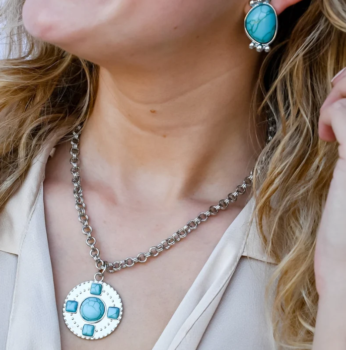 A close-up lifestyle shot of a woman wearing a silver-toned chain necklace with a round pendant featuring light blue stones, and a matching light blue stone earring.