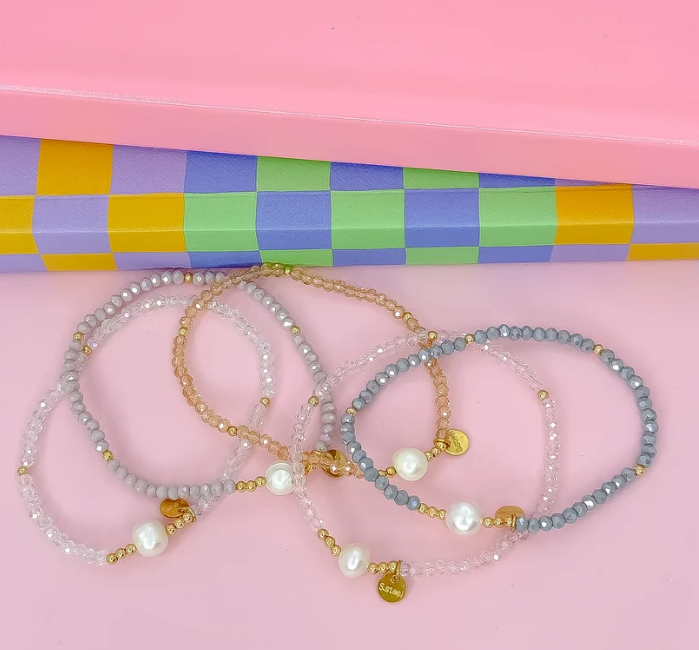 Four beaded stretch bracelets with gold-tone hardware and pearl-like beads, arranged on a pink surface in front of a pastel checkered box.