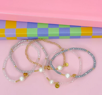 Four beaded stretch bracelets with gold-tone hardware and pearl-like beads, arranged on a pink surface in front of a pastel checkered box.