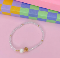 A beaded stretch bracelet featuring clear faceted beads, a white pearl, and small gold spacer beads, displayed on a pink surface with a colorful checkered gift box in the background.