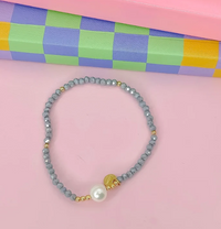 A beaded stretch bracelet featuring light blue-gray round beads, small gold spacer beads, a white faux pearl, and a gold crescent moon charm, displayed on a pink background with a colorful checkered box.