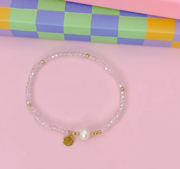 A delicate beaded bracelet with clear faceted beads, small gold spacer beads, a white pearl, and two gold charms, displayed on a pink background with a colorful checkered box in the background.