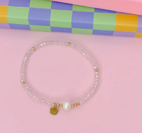 A delicate beaded bracelet with clear faceted beads, small gold spacer beads, a white pearl, and two gold charms, displayed on a pink background with a colorful checkered box in the background.