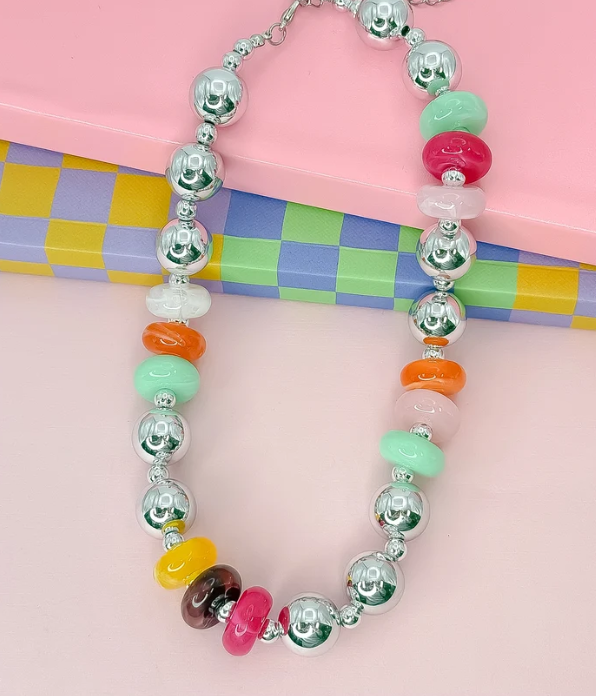 A beaded necklace with alternating shiny silver metallic beads and pastel-colored disc beads in pink, mint green, orange, yellow, and brown, displayed on a pink background with a checkered box.