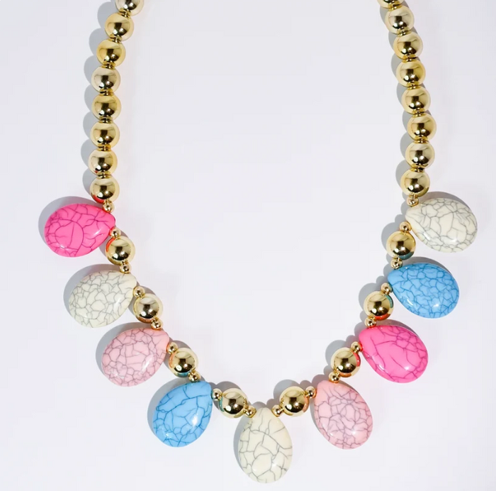 A statement necklace featuring a gold beaded chain and teardrop-shaped crackle enamel charms in pink, blue, and cream, arranged symmetrically on a white background.