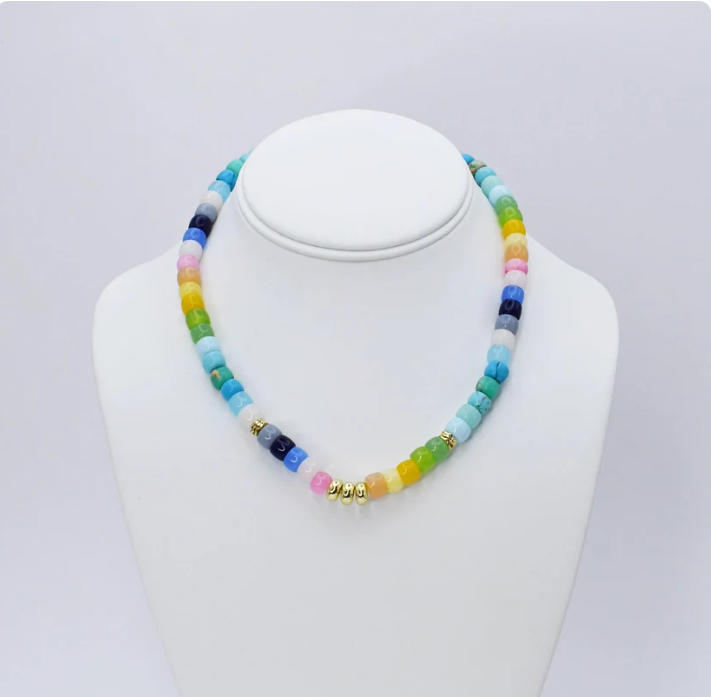 A beaded necklace displayed on a white jewelry bust against a light gray background. The necklace features multiple translucent round beads in rainbow colors including blue, green, yellow, pink, and orange, separated by small gold spacer beads.