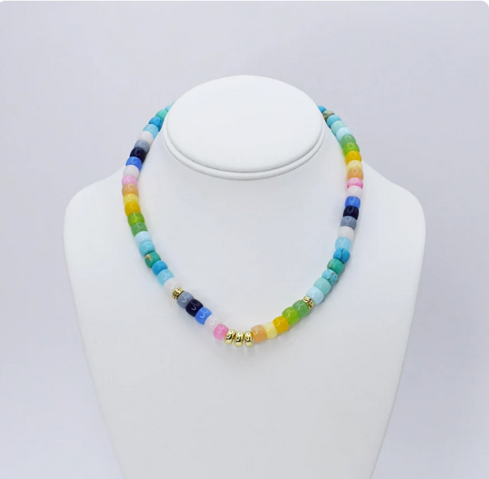 A beaded necklace displayed on a white jewelry bust against a light gray background. The necklace features multiple translucent round beads in rainbow colors including blue, green, yellow, pink, and orange, separated by small gold spacer beads.