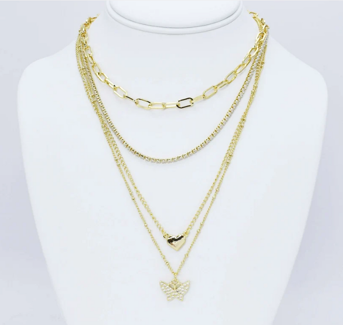 A gold multi-layered necklace set displayed on a white mannequin bust. The set includes a chunky paperclip chain, a fine curb chain, a delicate chain with a small heart pendant, and a longer chain with a butterfly pendant.