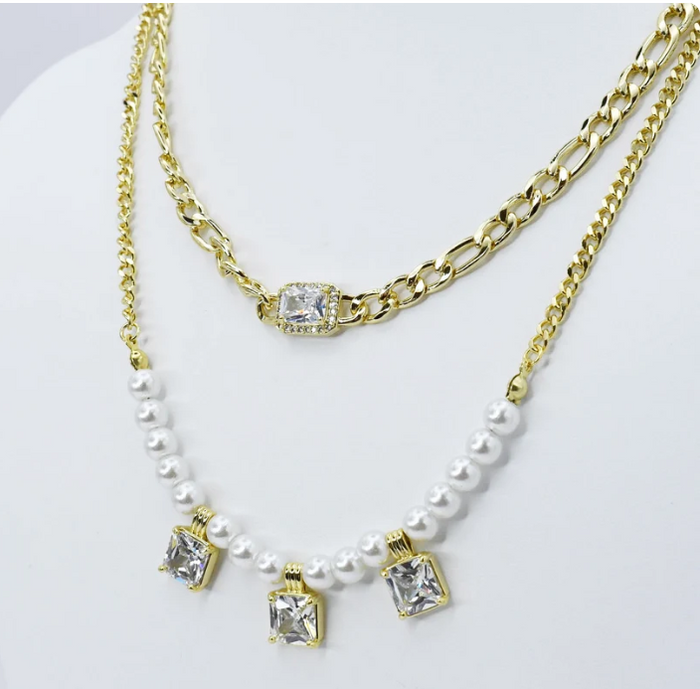 A double-layered gold necklace set displayed on a white mannequin bust. The top layer features a gold Figaro chain with a rectangular clear rhinestone pendant.