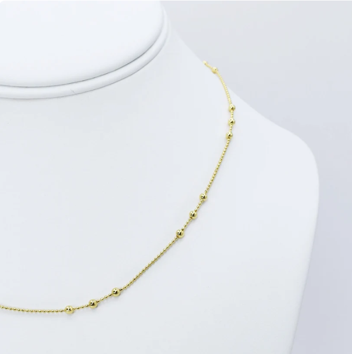 A delicate gold chain necklace with small bead accents, displayed on a white jewelry bust against a light gray background.