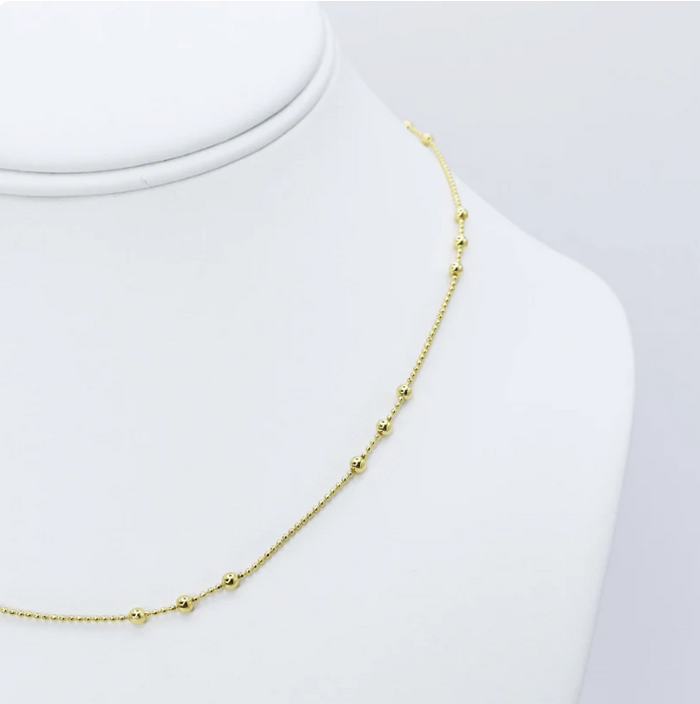 A delicate gold chain necklace with small bead accents, displayed on a white jewelry bust against a light gray background.