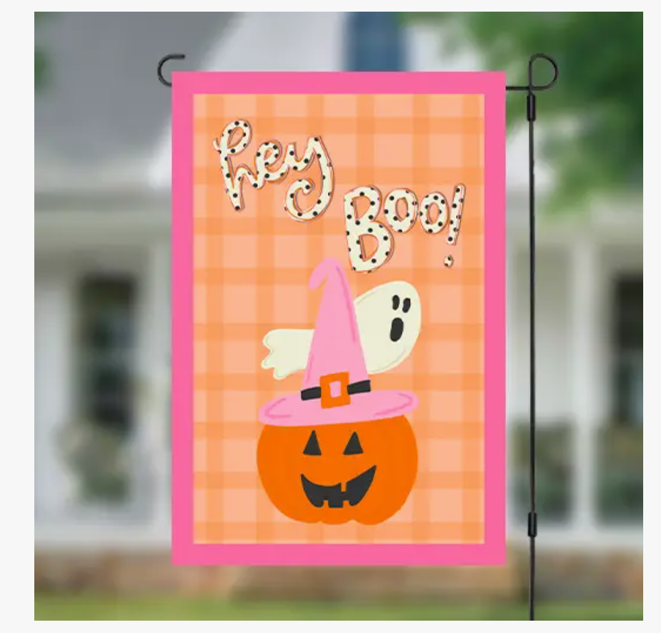 A Halloween garden flag hanging on a black metal stand, featuring a pink border, orange plaid background, a smiling jack-o'-lantern, a white ghost, and the words 'Hey Boo!' in a dotted font. The background shows a blurred outdoor scene of a house and greenery.