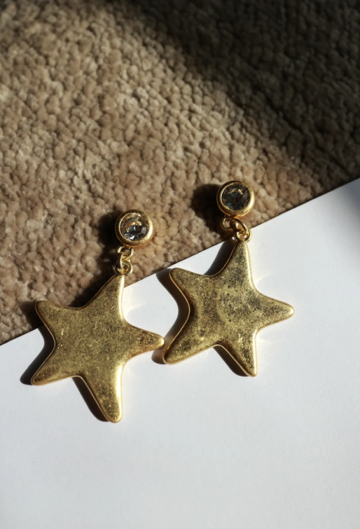 A pair of gold star-shaped dangle earrings with small clear stone studs, displayed on a white surface with a textured beige rug in the background.
