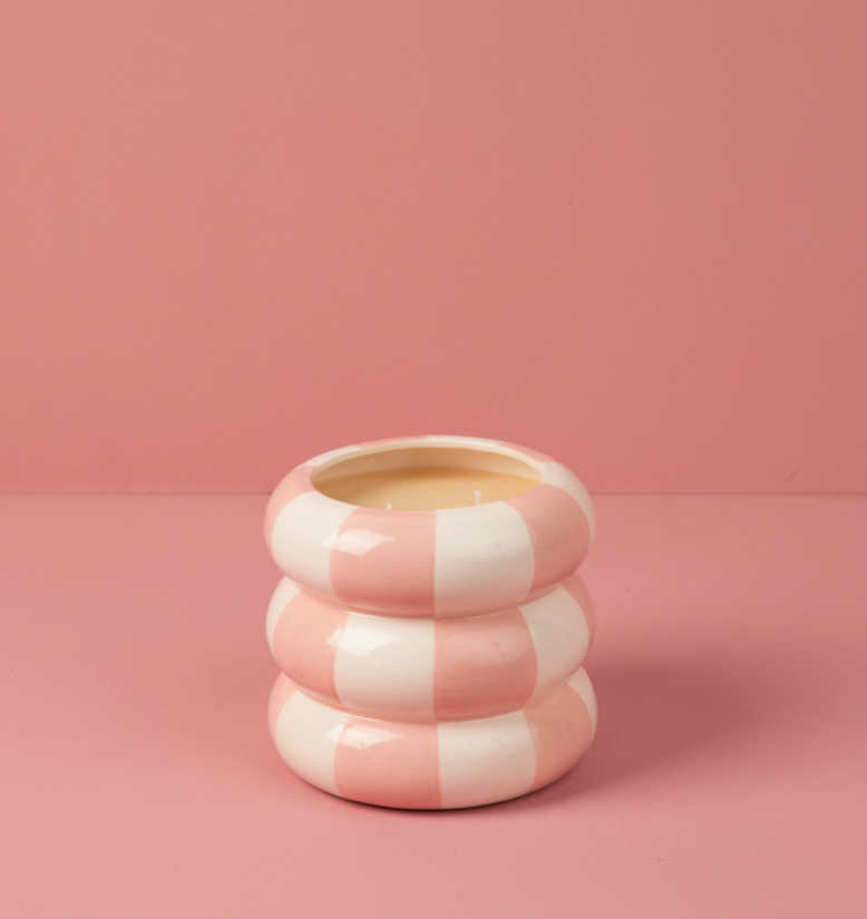 A pink and white striped ceramic candle holder with a rounded, stacked design, holding a yellow candle, set against a solid pink background.