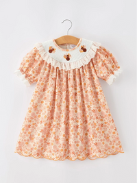 A short-sleeve baby dress with a peach and orange floral print, featuring a white ruffled Peter Pan collar decorated with three embroidered turkeys hanging on a wooden hanger against a light gray background.
