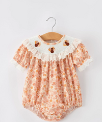 A baby girl's short-sleeve romper in a peach and orange floral print, featuring a white ruffled collar with embroidered cartoon birds and lace trim, hanging on a wooden hanger against a light gray background.