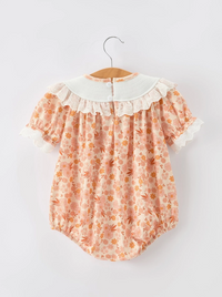 A baby romper with a peach and orange floral print, featuring a white ruffled collar and lace trim, hanging on a wooden hanger against a light gray background.
