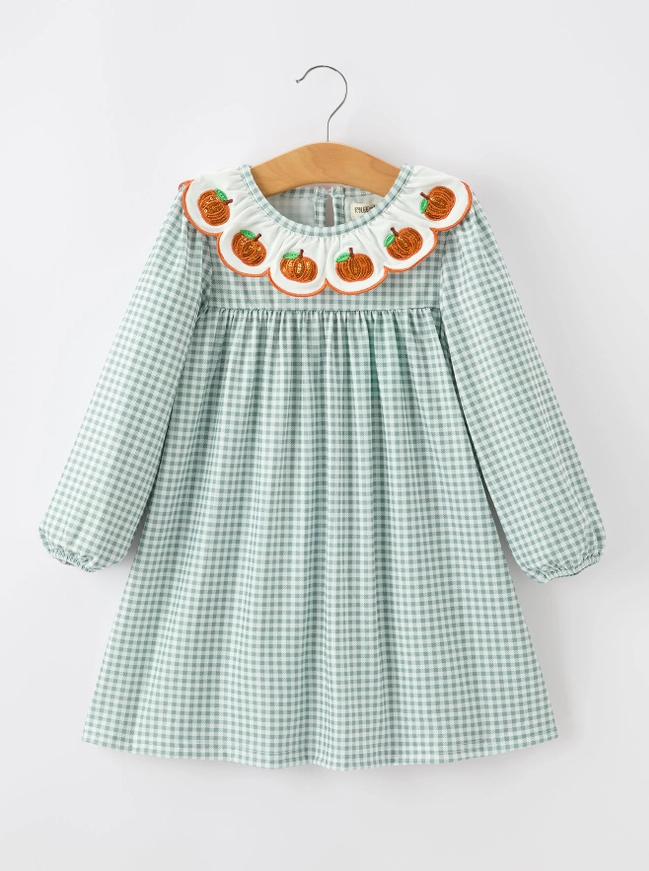 A toddler girl's long-sleeve dress in a light green and white gingham pattern, featuring a decorative scalloped collar with embroidered orange pumpkins and white ruffles. The dress is shown hanging on a wooden hanger against a plain white background.