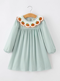 A toddler girl's long-sleeve dress in a light green and white gingham pattern, featuring a decorative scalloped collar with embroidered orange pumpkins and white ruffles. The dress is shown hanging on a wooden hanger against a plain white background.