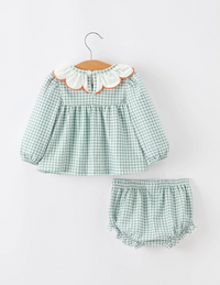 A two-piece baby girl's outfit set featuring a long-sleeve green and white gingham dress with a white ruffled collar trimmed in orange, paired with matching green and white gingham bloomers, displayed on a wooden hanger against a light gray background.