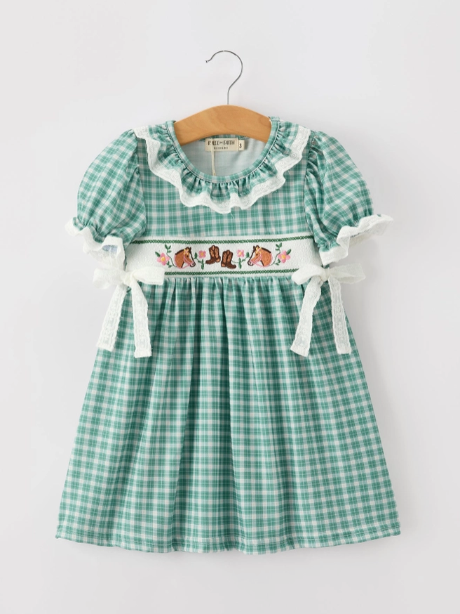 A green and white gingham baby girl's dress hanging on a wooden hanger against a light gray background. The dress features a ruffled lace collar, short puff sleeves with white lace ties, and a smocked waistband decorated with a floral and horse embroidery 
