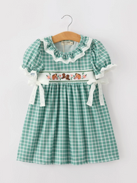 A green and white gingham baby girl's dress hanging on a wooden hanger against a light gray background. The dress features a ruffled lace collar, short puff sleeves with white lace ties, and a smocked waistband decorated with a floral and horse embroidery 