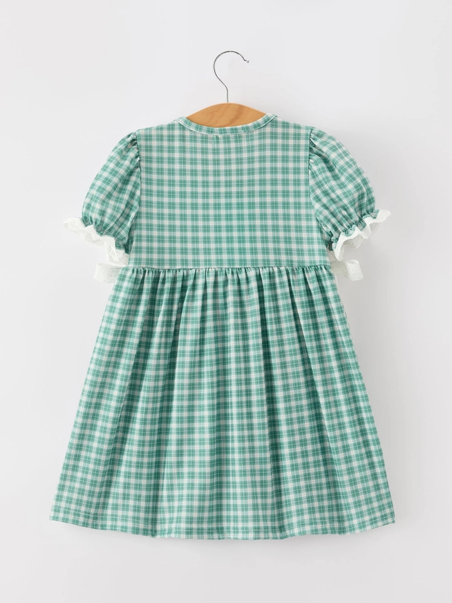 A green and white gingham check dress with short puff sleeves and white ruffle trim, hanging on a wooden hanger against a plain light gray background.